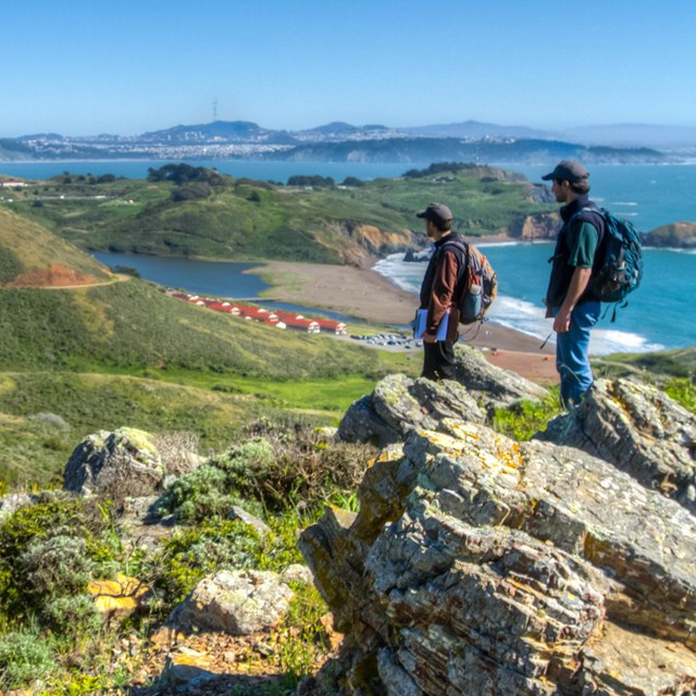two people in bakcpacks look out at a view from the top of a steep trail.