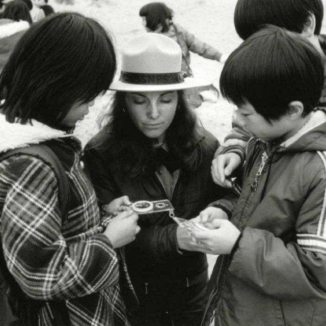 A black and white photo of a ranger looks at a hand lens with two youths. 