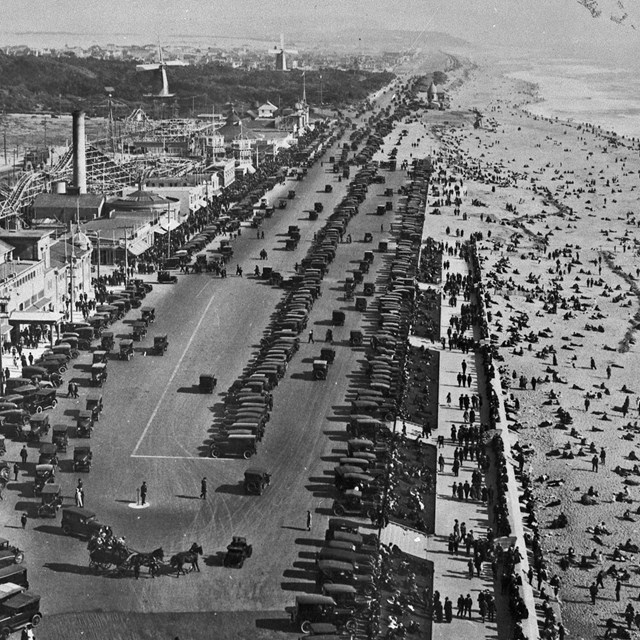 Historic photo of a crowded Ocean Beach and parking lots full of 1920s cars  