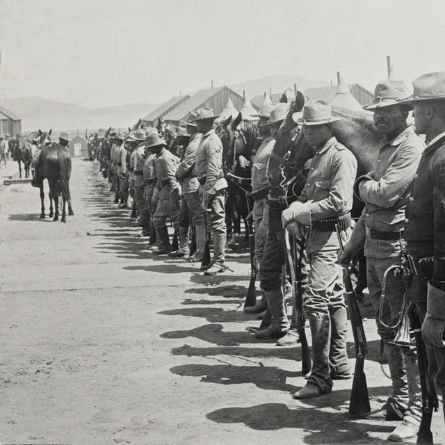 A group of Black cavalry soldiers and their horses stand outside.