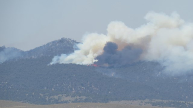 Smoke billows from a dark tree covered hillside in the distance. Flames are visible near the center.