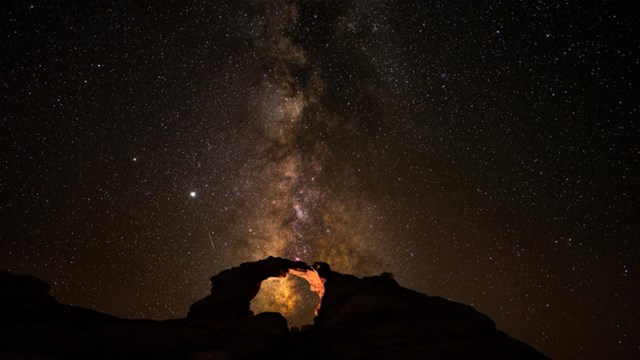 A color night photo of a natural, slightly lit arch with the milky way stretching above it.