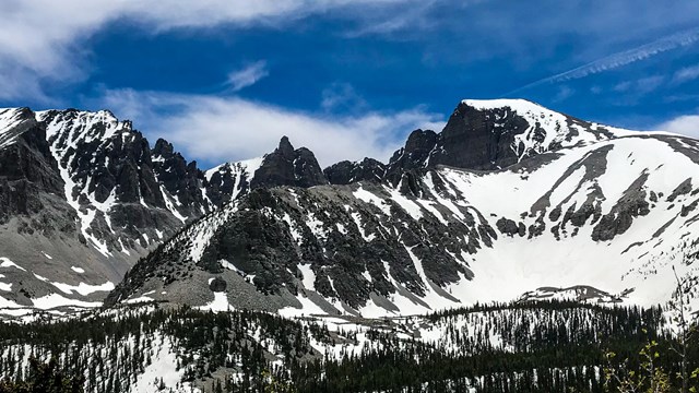 Snow covered gray cliffs and mountains stand in the distance under a patchy blue sky
