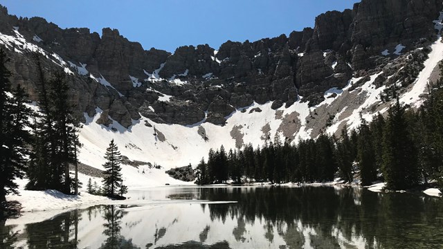 An alpine lake surrounded by tall pine trees is overlooked by sheer grey cliffs with patchy snow