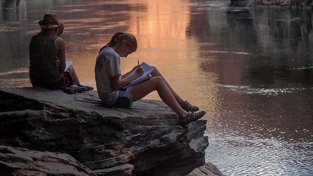 Two people sit by the Colorado River in the Grand Canyon working on a workbook