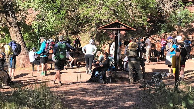 A large group gathers at a rest area and are donning hiking attire. 