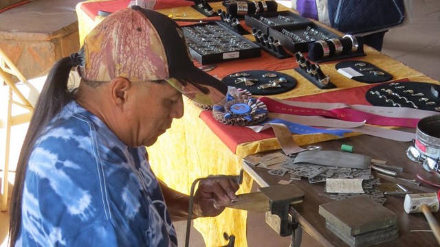 A man sits at a table with silver works and a display of silver jewelry 