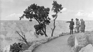 black and white photo from 1930's of park ranger escorting well dressed visitors on rim trail.