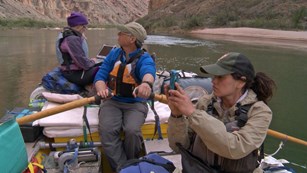 Three scientists in a raft on a green river. One is photographing, one is rowing, one with a laptop.