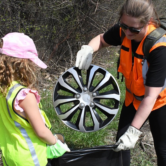 A woman in an orange vest is working with a young girl to bag a tire rim of Earth Day.