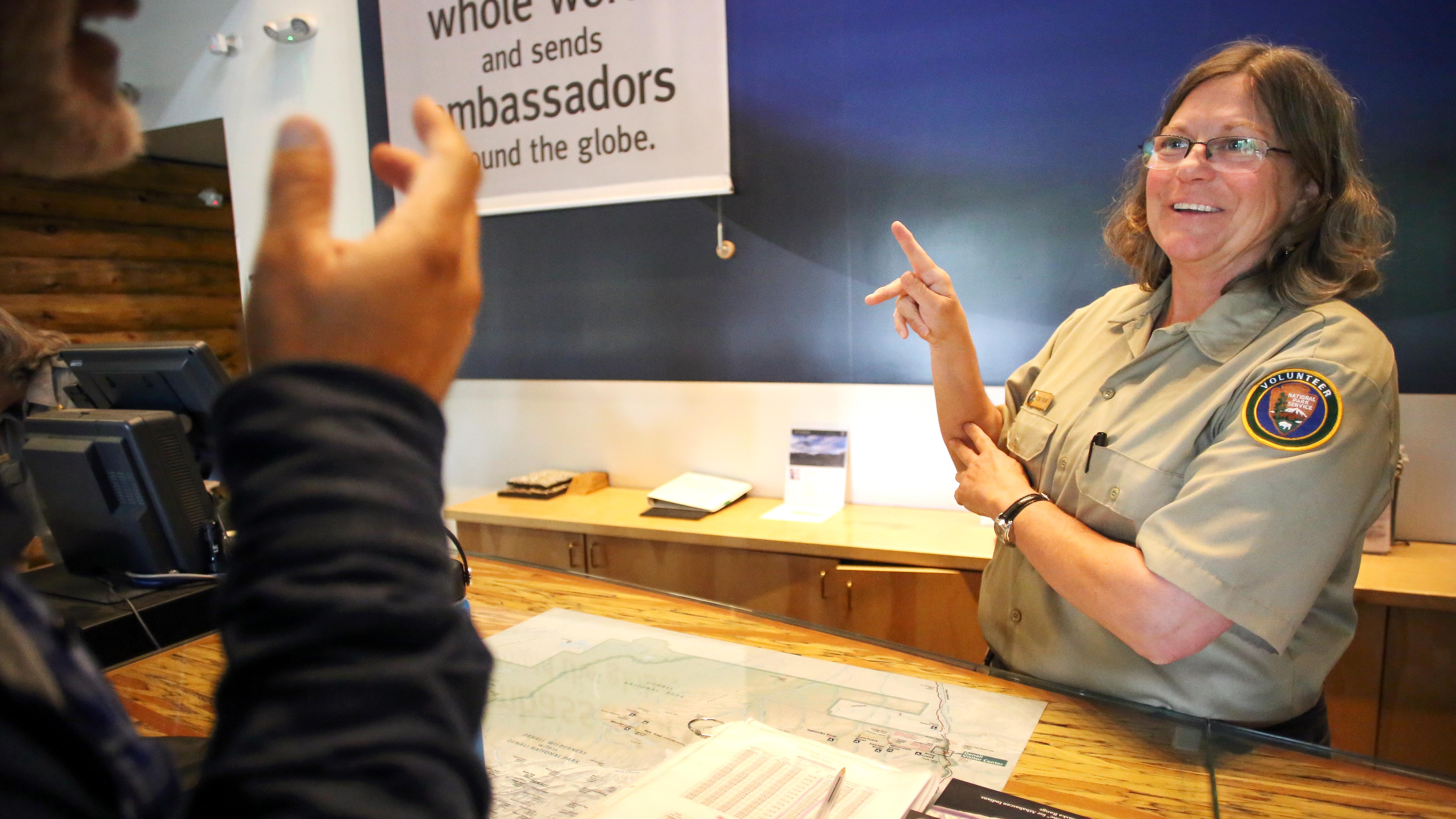 A volunteer behind a desk communicates using sign language with a visitor.