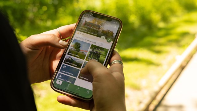 A person holds a cell phone in their hand in front of a trail, on the NPS app!