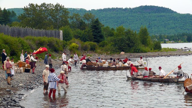 Several canoes carrying people in historic clothing land on a beach.