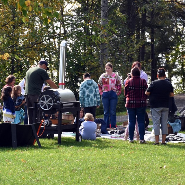 People standing outdoors around a metal machine next to a birch lodge.