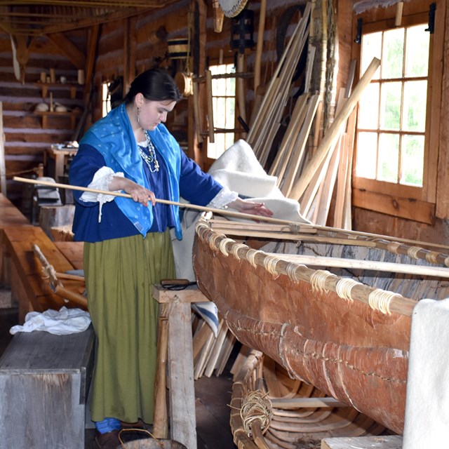 A person in historic clothing measuring a length of wood for a canoe rib.