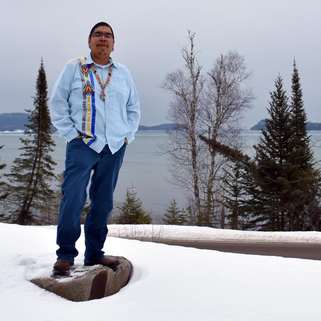 A person wearing a traditional Ojibwe ribbon shirt, standing on a rock in snow in front of a lake.