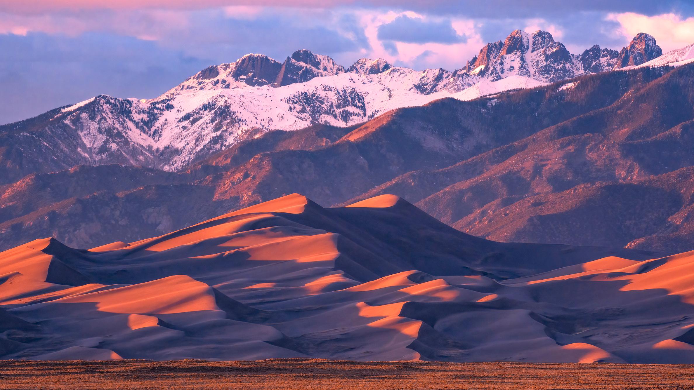 Orange light on Sand dunes with Snow capped peaks in background, with grasses in foreground