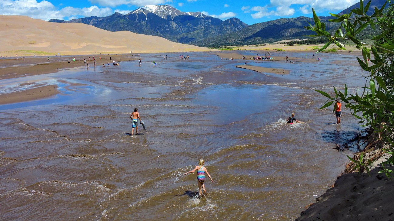 A wide creek with people playing in it at the base of dunes and a snow-capped mountain