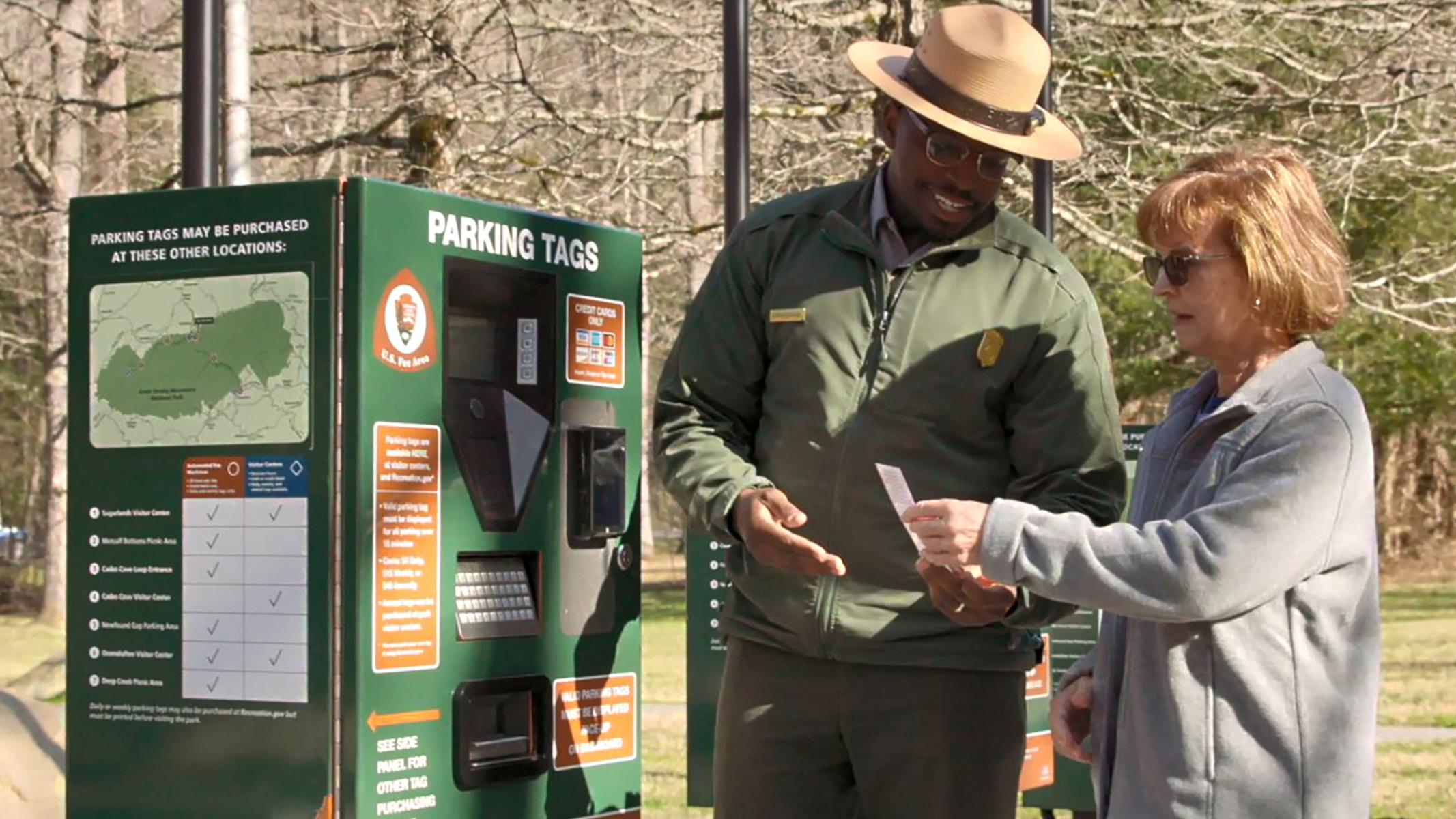 A person handing a parking tag to another person beside a green parking tag kiosk.