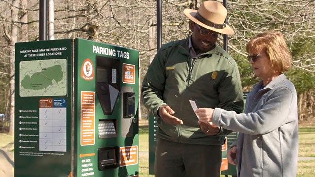 A person handing a parking tag to another person beside a green parking tag kiosk.