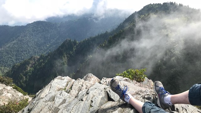 A hiker's feet resting on a rock at a mountain overlook with some fog and clouds.