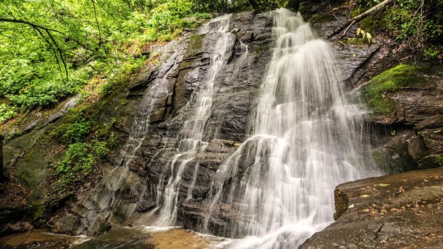 A waterfall cascading down rocks, some with moss and greenery. Green trees surround the falls.