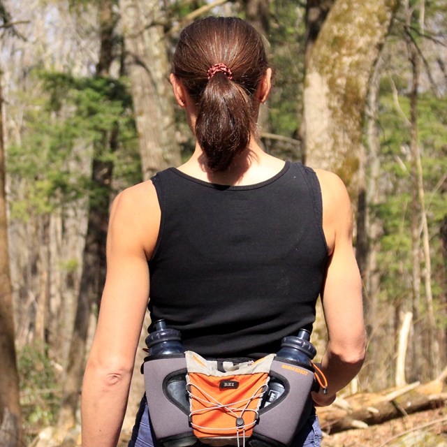 The back of a hiker walking down a trail with water strapped to her waist on a waist band.