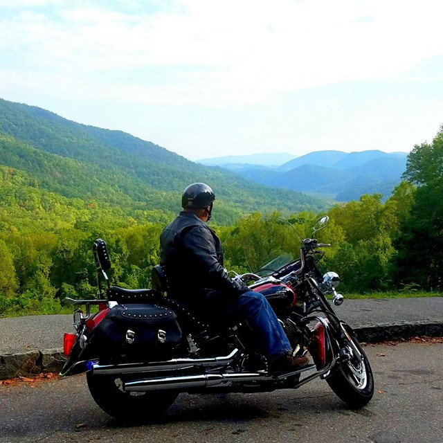 A motorcyclist at a scenic mountain pull out, wearing a leather jacker, pants, and a black helmet.