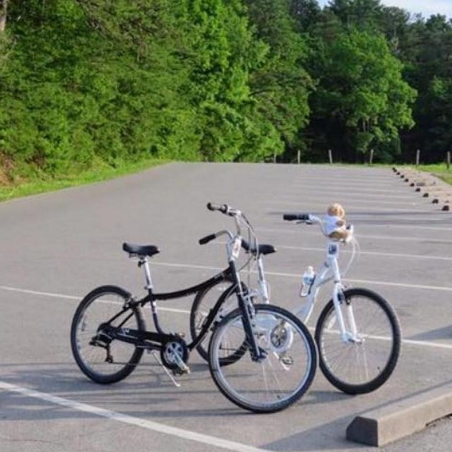 Two bikes in a paved parking lot. Green trees in the background.