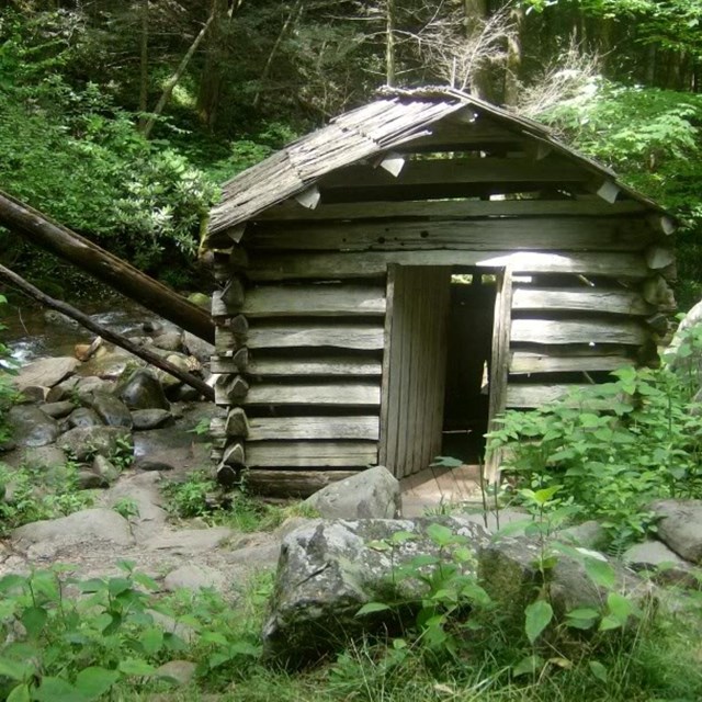 A small, rustic, faded log cabin near a creek surrounded by rocks and greenery