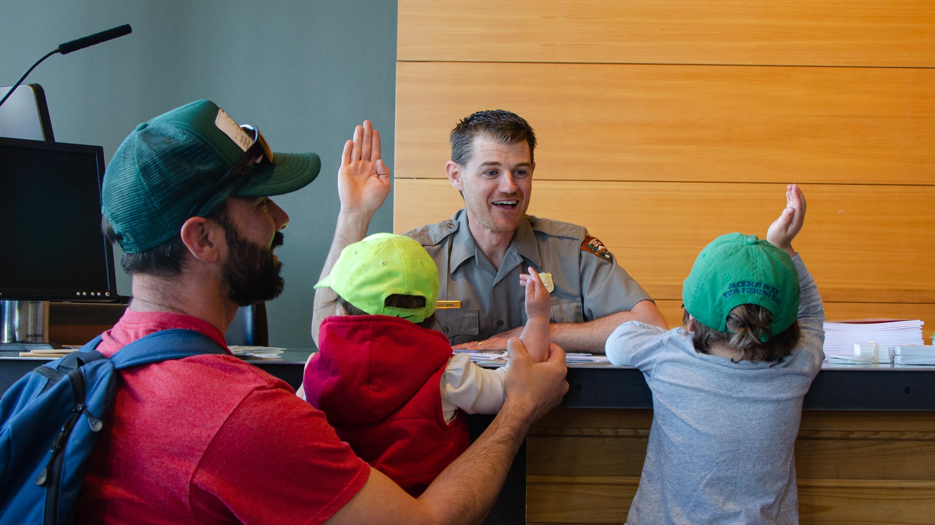Two kids are sworn in as Junior Rangers by a park ranger at a visitor center front desk.