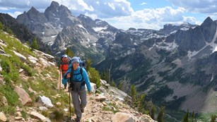 Two backpackers climbing a trail in the Teton Range with the Grand Teton in the distance.
