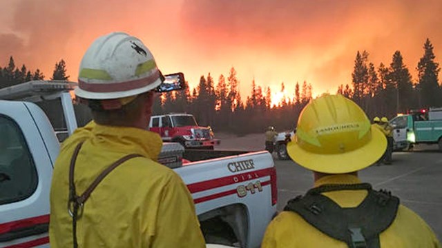 Two firefighters stand in a large parking lot, while a fire approaches from beyond the trees.