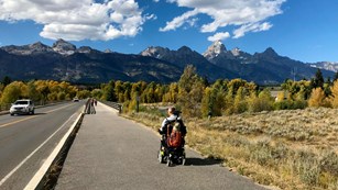 A visitor in a wheelchair on a paved path with mountains in the background.