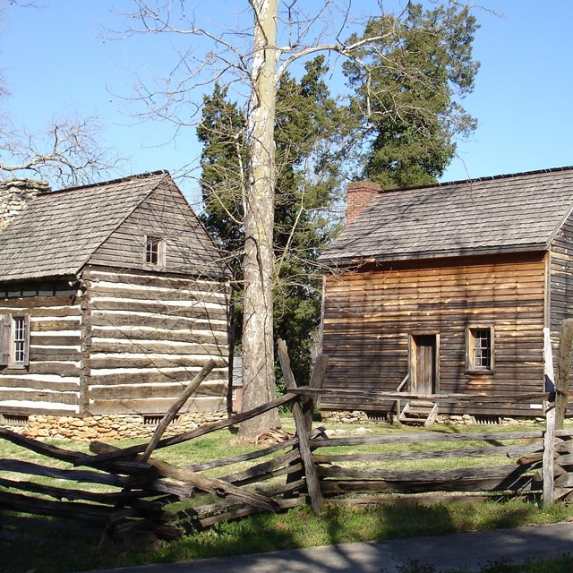 Two colonial buildings stand behind a split rail fence, with a tree in the middle