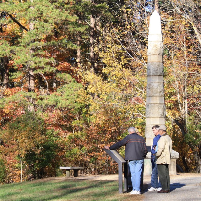Three men read an exhibit panel next to an obelisk monument, forest behind