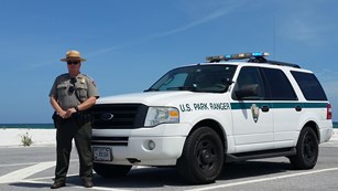 A park ranger stands in front of a patrol SUV just in front of a beach.