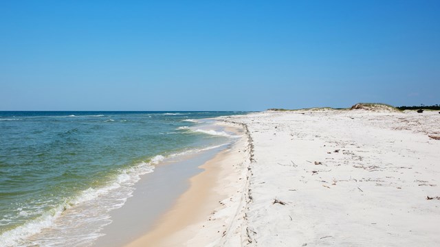 Small waves crash from left to right on to a barrier island with vegetated dunes to the right.