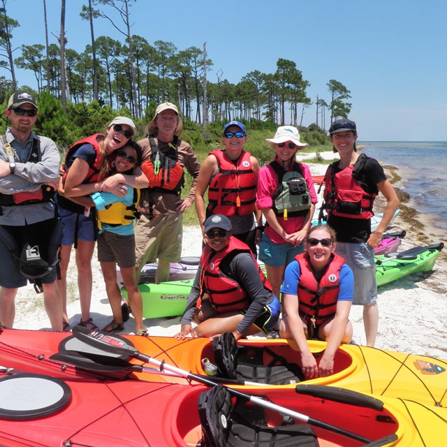 A group of young adults in life-jackets gather around kayaks.