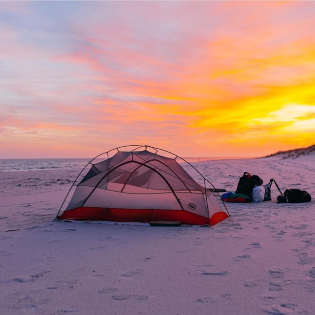A tent is set up on a beach with the sun setting in the background.