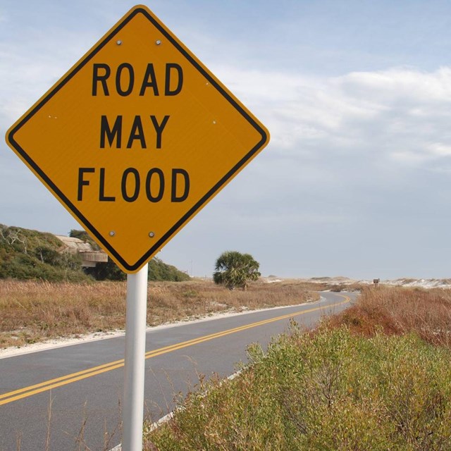 A sign reading road may flood stands next to a road along a beach.