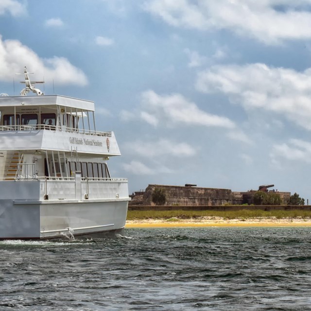 A large white tour boat just off shore of an island with a masonry fort in the background.