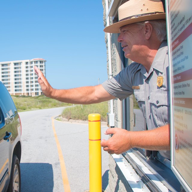 A ranger waves to visitors in a car as they enter a park.