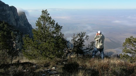 A hiker uses binoculars to view a desert mountain landscape.