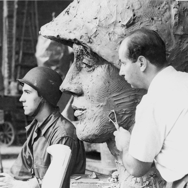 A young man poses next to a giant plaster sculpture of his head.