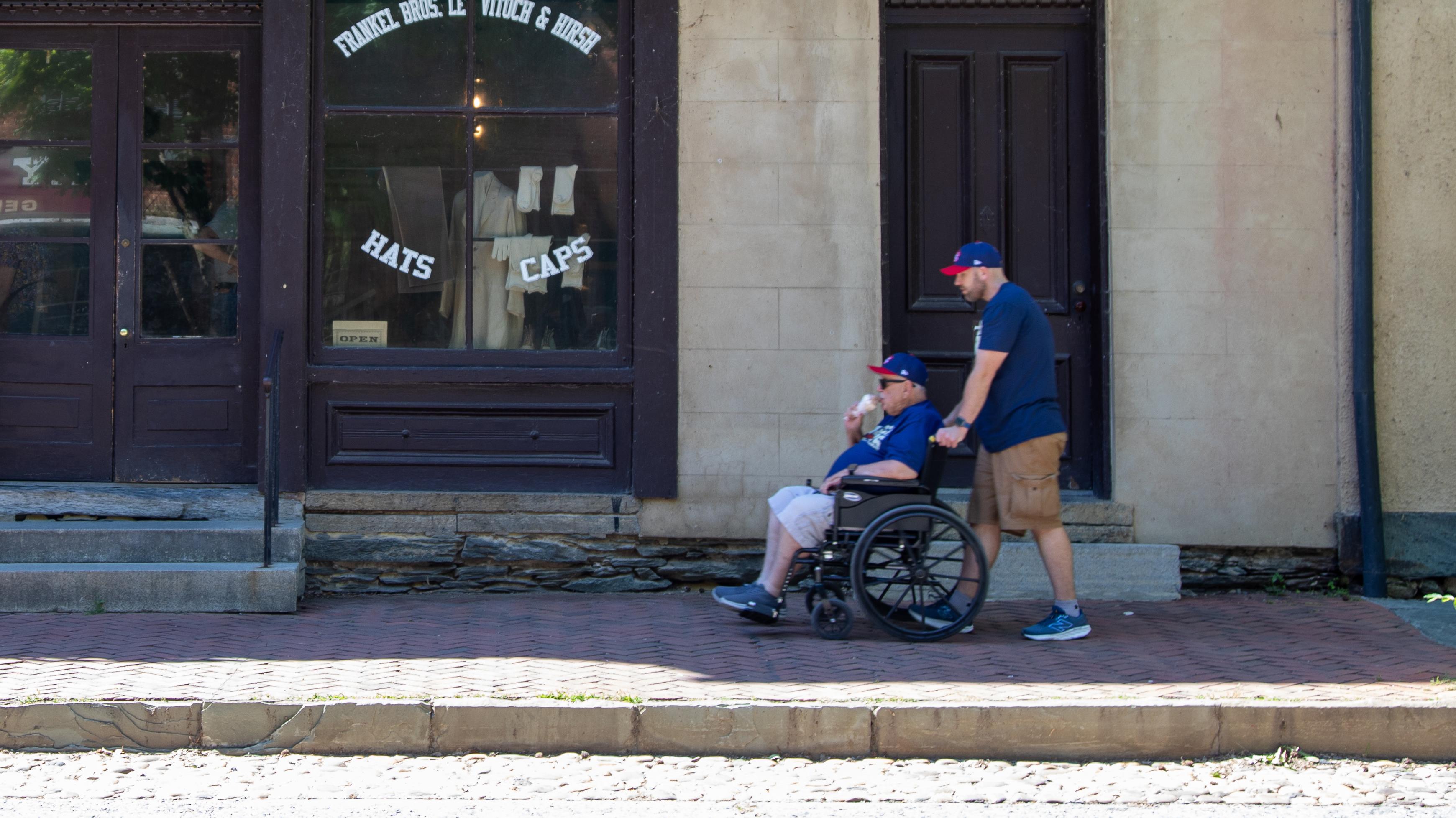A man pushes a man in a wheelchair on a brick sidewalk in front of a building.