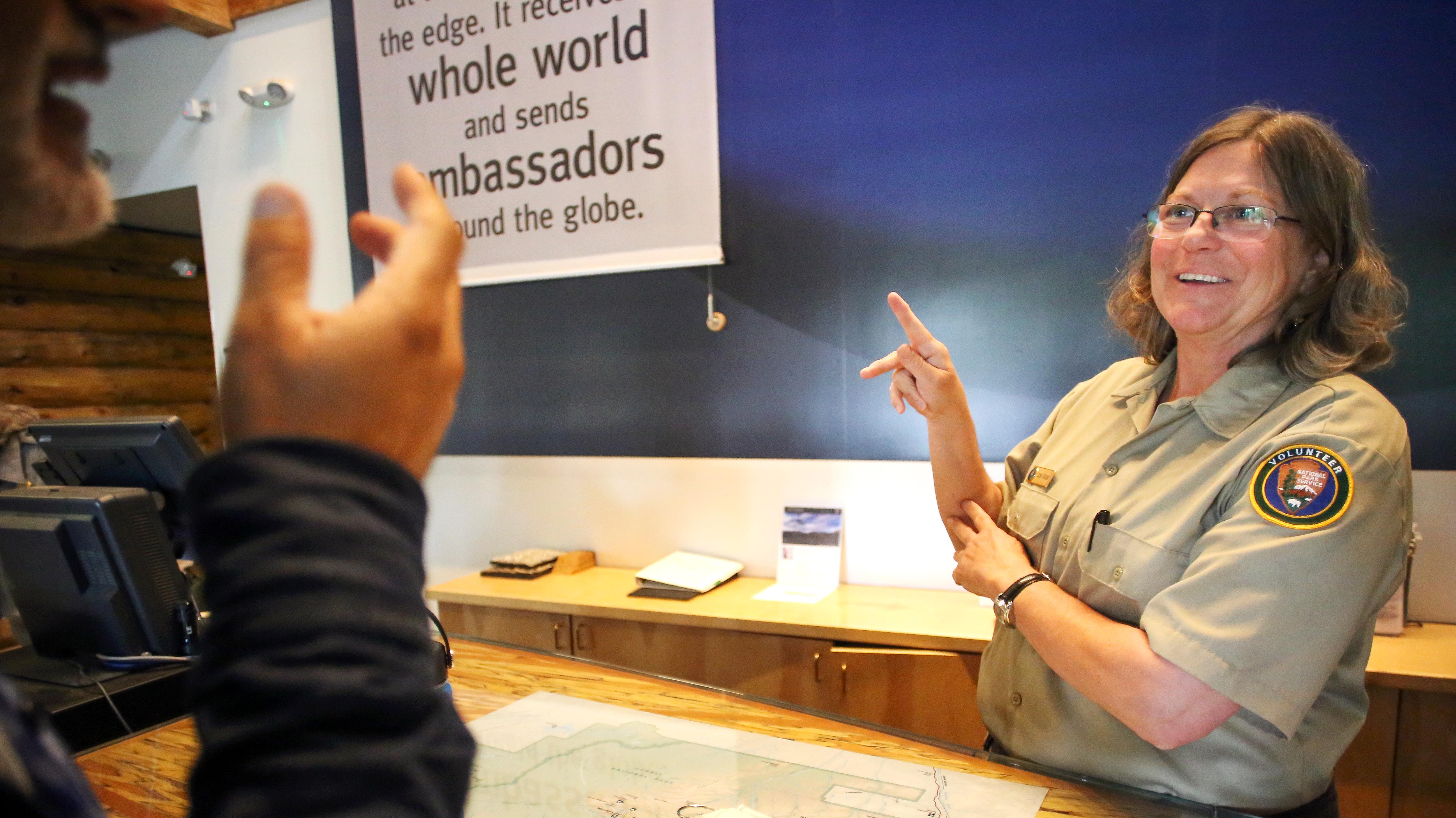 A park volunteer behind a desk communicates with a visitor using sign language.
