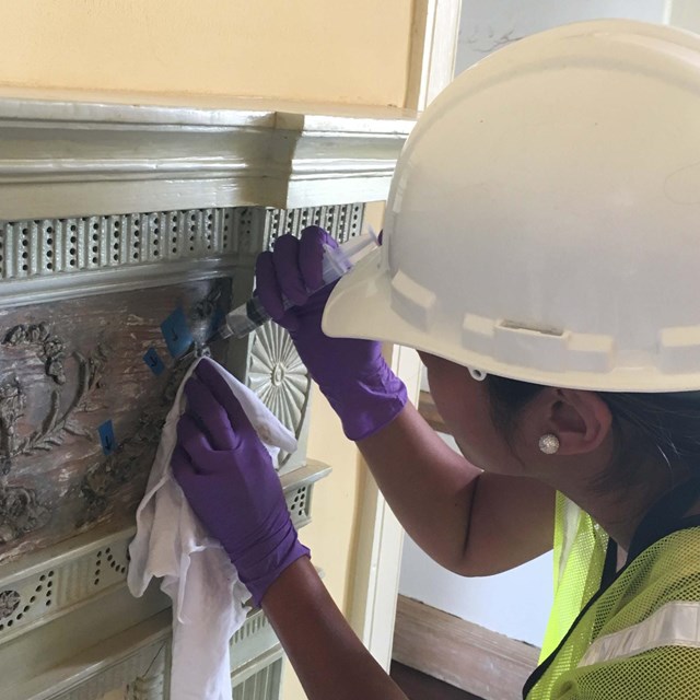 A woman in a hardhat makes repairs to an ornate wooden mantelpiece