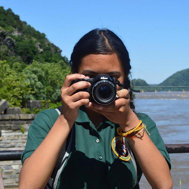 Dark-haired girl in park volunteer shirt holds camera toward photographer