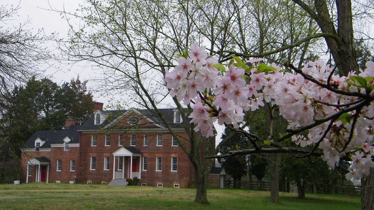 A red brick manor house sitting on a field of green grass. Trees are visible in the foreground. 